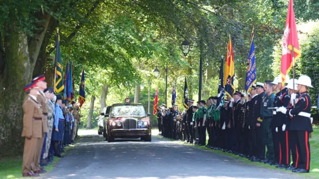 Military personnel in uniform and holding flags line a route as the King's motorcade drives past
