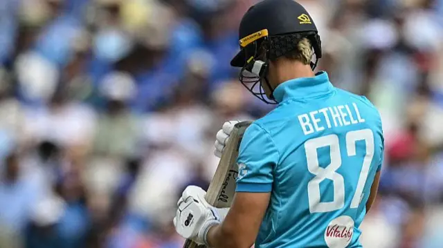 Jacob Bethell, facing away from the camera in a bright blue England shirt (with Bethell 87 on the back), dark blue helmet, and white gloves, inspects the end of his bat