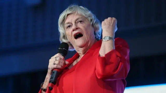 Ann Widdecombe wears a red dress and has short grey hair, she stands on a stage speaking into a microphone.