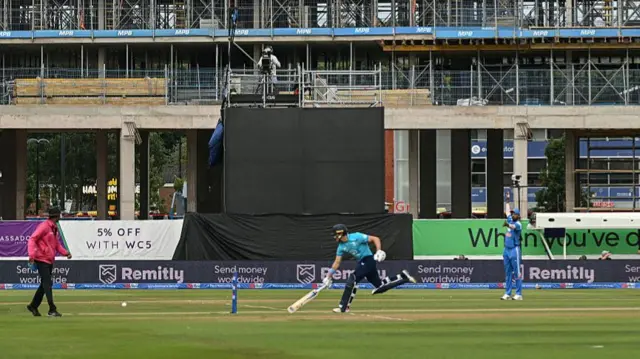 Jacob Bethell (centre), wearing a bright blue shirt, dark blue trousers and helmet, and white gloves, runs to slide his bat in at the non-striker's end