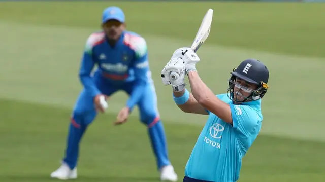 Jacob Bethell (right), wearing a bright cyan blue England shirt and dark blue helmet, plays a shot with his bat high above his right shoulder, with an India fielder, wearing all blue, out of focus in the background