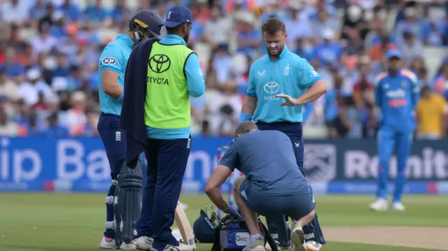Ben Duckett, right, wearing a bright blue England shirt, looks down at his injured left hand, with two players stood to his left and a physio, wearing a dark blue t-shirt and shorts, crouched down in front of him