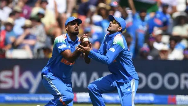 Gurnoor Brar (left) and Shubman Gill (right), both wearing blue kits and blue caps, collide with the white ball in between them in Brar's hands