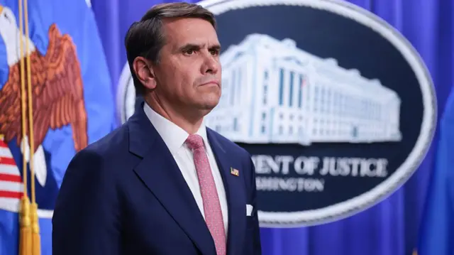 Todd Blanche stands inside the Department of Justice. He wears a blue suit and pink tie