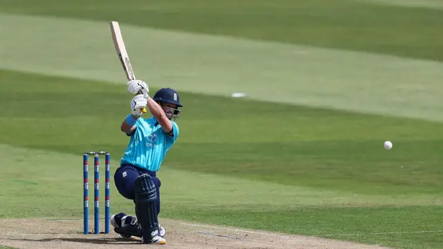 Ben Duckett, wearing a bright blue England shirt, dark blue trousers and helmet, down on one knee with his bat held high in two hands above his right shoulder, with the white ball flying away (right)