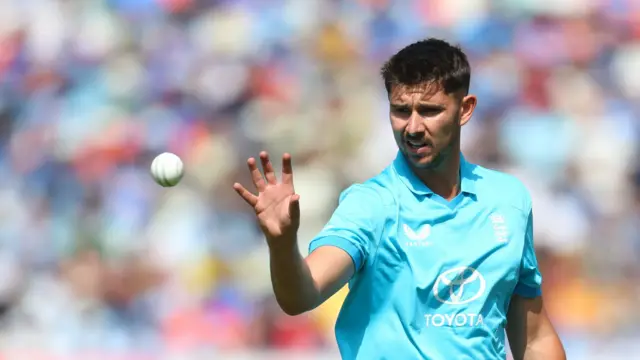 Josh Tongue, wearing a bright blue England shirt, holds up his right hand to catch the white ball