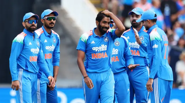 Jasprit Bumrah, wearing an all blue India kit, runs his left hand through his hair and smiles with his teammates around him