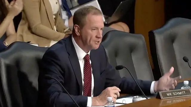 Eric Shmitt wears a navy suit and burgundy tie and sits at a wooden table in front of a microphone