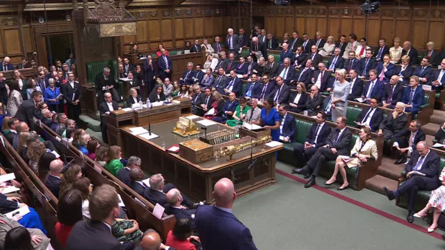 The House of Commons, with Kemi Badenoch standing up in a blue dress among MPs