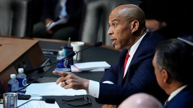 Senator Cory Booker is pictured behind the senate judiciary committee bench, wearing a suit and tie, and gesturing with his left hand.
