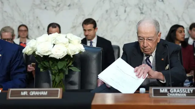 A bouquet of white roses sits on a committee table behind a name card reading "Mr Graham". To the right, an older man sits behind a name card reading "Mr Grassley, Chairman".