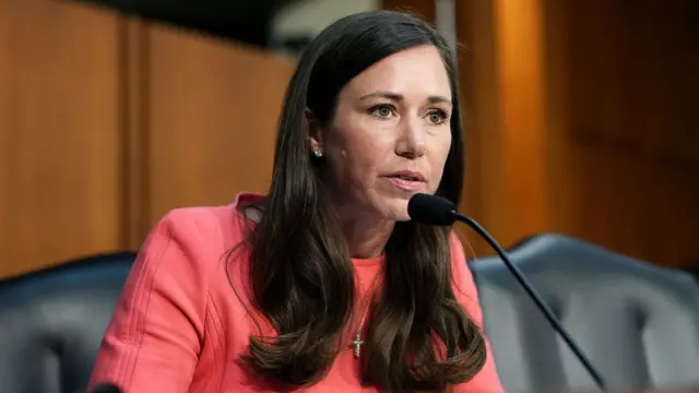 Katie Britt is pictured speaking in a senate committee room, wearing a salmon pink top.