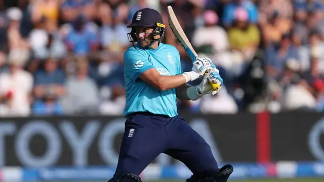 Joe Root, wearing a bright blue England shirt, dark blue trousers and helmet, holds his bat in two hands by his left shoulder and looks behind him over his right