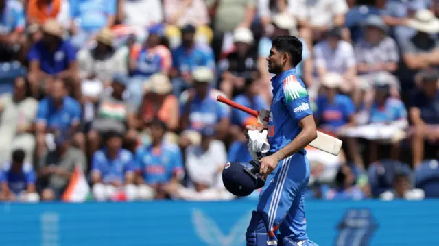 Shubman Gill, wearing all blue with his dark blue helmet in his left hand and bat tucked under his right arm, leaves the field in front of a blurred crowd full of India shirts