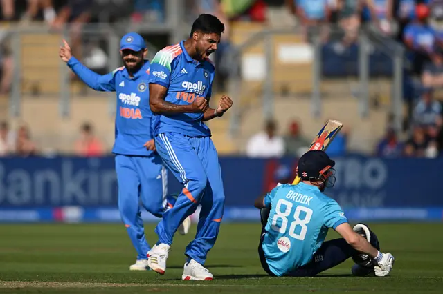 Gurnoor Brar (centre), wearing all blue, roars and clenches his fists with dismisses batter Harry Brook, wearing a blue England shirt and dark blue helmet facing away from camera, sat at his feet