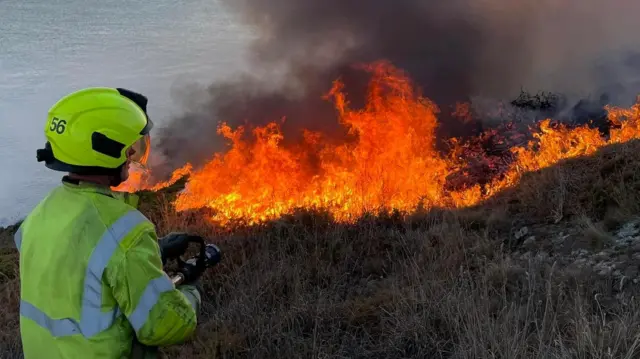 A firefighter in protective clothing standing at the edge of a moorland wildfire by the sea and using a swatter to try and extinguish the flames. Gorse and vegetation is ablaze in the background.
