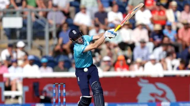 Harry Brook, wearing a bright blue England shirt and dark blue trousers and helmet, with his bat high to his left is struck in the midriff by the white ball
