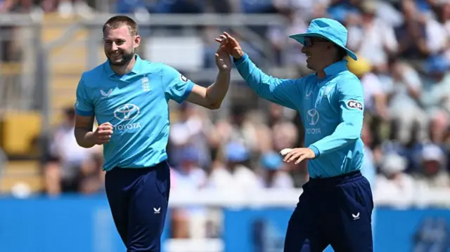 Gus Atkinson (left), wearing a bright blue shirt and dark trousers, high fives Harry Brook (same outfit, with wide-brimmed bright blue hat) to his left
