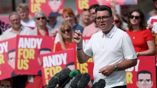 Andy Burnham speaks delivers a speech in front of his supporters, some holding 'Andy for us' signs. Burnham is standing in front of a range of microphones and is wearing a white polo neck