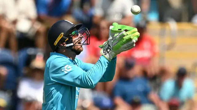 Jos Buttler, wearing a bright blue England shirt, dark blue helmet and sunglasses, holds up his green gloves to catch the white ball