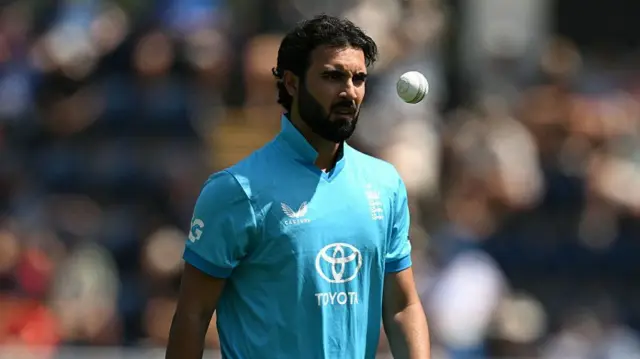 Saqib Mahmood, wearing a bright blue England shirt, tossing the white ball up in front of his eyes