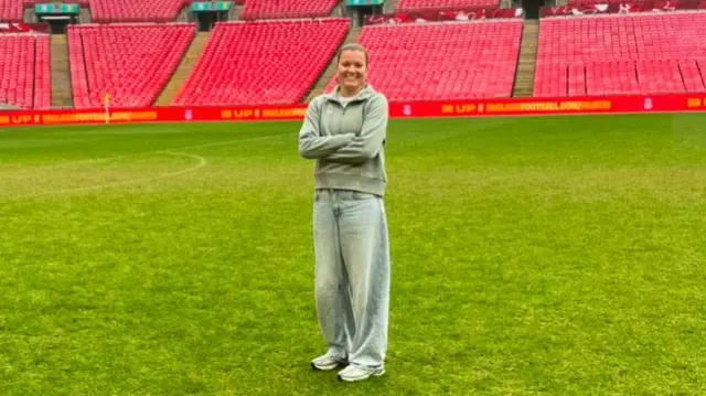 Beatrice Rogers, a young white woman with dark hair pulled back in a pony tail, wearing a quarter zip fleece, jeans and trainers standing on a large green football pitch with red seats behind.
