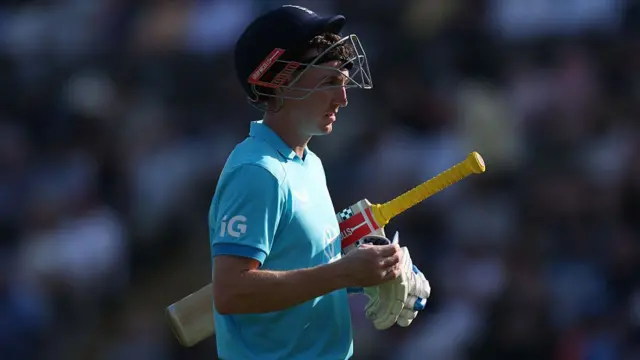 Harry Brook, wearing a bright blue England shirt, walks off the field holding his gloves, with his bat tucked under his left arm and his dark blue helmet tipped back on his head