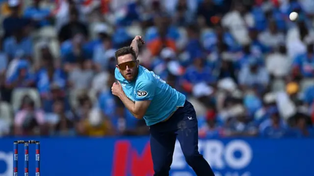 Liam Dawson, wearing a bright blue England shirt, dark blue trousers and glasses, bowling