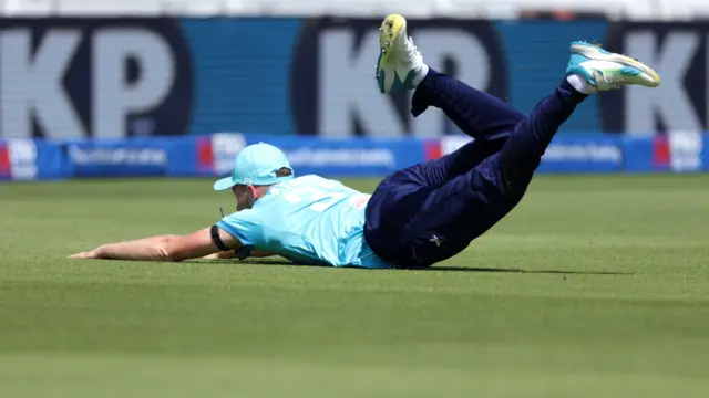 Gus Atkinson, diving away from the camera with his arms outstretched across the floor and legs in the air after dropping a catch, wearing a bright blue cap and shirt and dark blue trousers