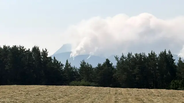 smoke emerging from a hill in the distnace, behind some trees and a field