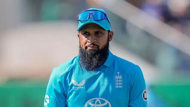 Head and shoulders of Adil Rashid, wearing a bright blue England shirt and cap with sunglasses on the peak