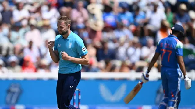 Gus Atkinson (left), wearing a bright blue England shirt and dark trousers, clenches his fists and smiles with Shubman Gill (right), wearing blue, walking off in the opposite direction