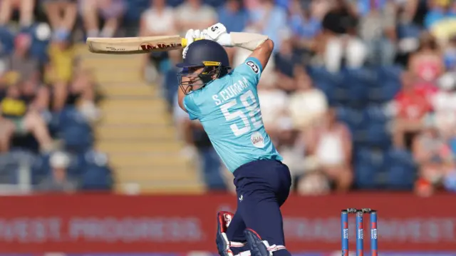 Sam Curran, playing in a bright blue England shirt - with S Curran 58 in white on the back - and dark blue trousers, helmets - follows through a drive, with his bat parallel to the ground above his head