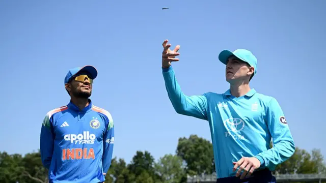 Harry Brook (right), wearing a bright blue England shirt and cap, tosses a coin in the air with his right hand, with India captain Shubman Gill, wearing a blue shirt, cap and sunglasses, watching on