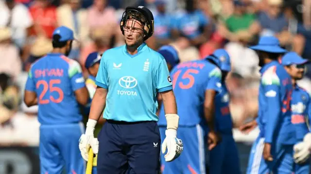Joe Root, wearing a bright blue England shirt, dark blue trousers, and white gloves, stands with his bat by his side in his right hand and his dark blue helmet pushed back on his head