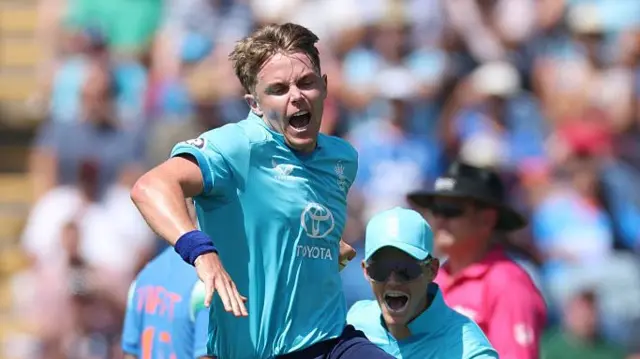 Sam Curran, wearing a bright blue England shirt, leaping in celebration with his mouth open