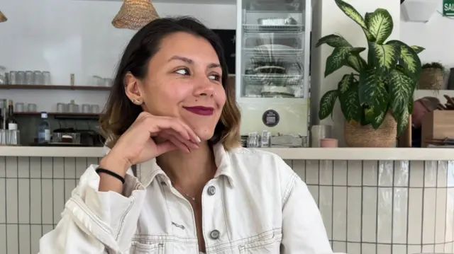 Micaela Basilico wearing a white denim-style jacket in a cafe, looking to the side and smiling.