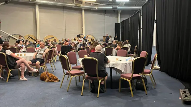 A roof laid out with large round tables, surrounded by chairs. They are being occupied by people in summer clothes, looking dejected and bored.