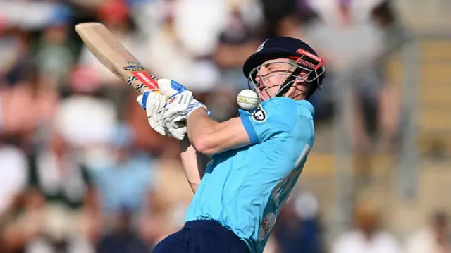 Harry Brook, wearing a bright blue England shirt and dark blue helmet, is struck on the grille by the white ball