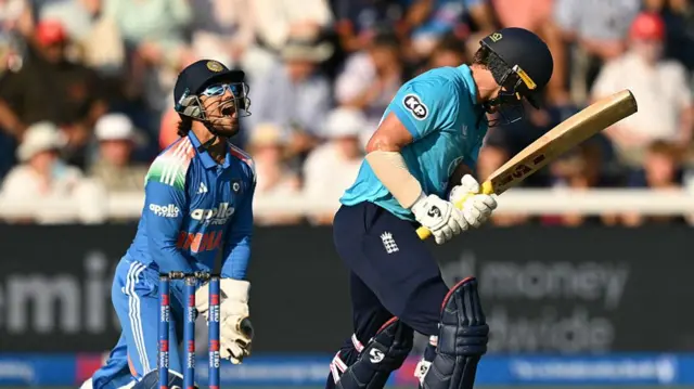 India's Ishan Kishan (left, wearing all blue, a dark blue helmet, sunglasses and white gloves) reacts after catching the ball to take the wicket of England's Sam Curran, wearing a bright blue shirt, dark blue trousers, and looking back at the wicketkeeper