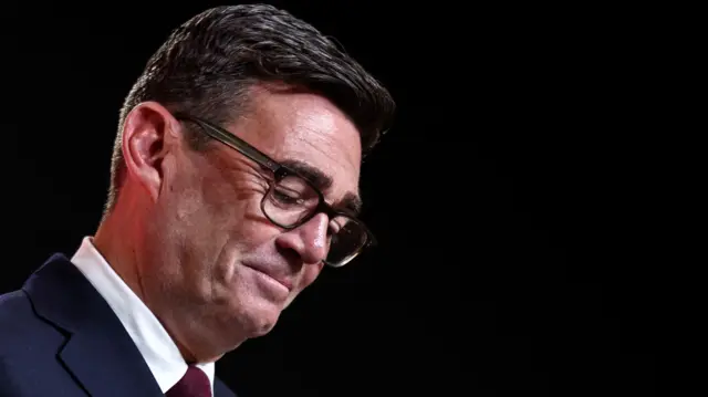 Andy Burnham wears a blue suit and burgundy tie and black glasses