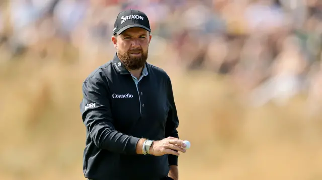 Shane Lowry walking in a black shirt and cap holding a golf ball.