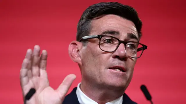 Close up of Labour leader Andy Burnham with his right hand raised as he speaks into a thin black mic. There's a red wall behind him