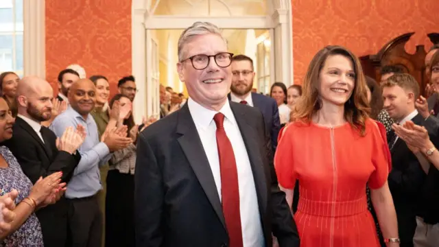 Keir Starmer and his wife Victoria surrounded by several people clapping.