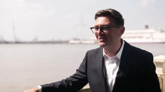 Andy Burnham stands by a waterfront wearing a black suit and white shirt