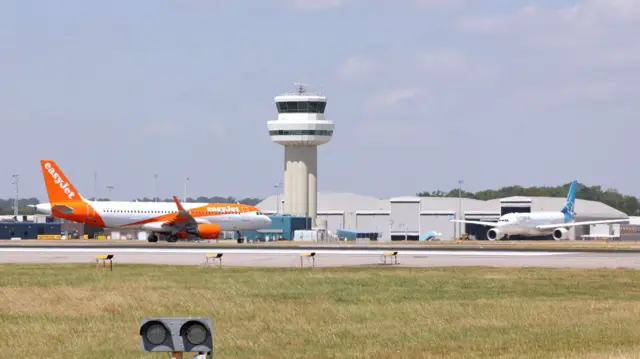 An EasyJet plane with white and orange livery taxis past the Gatwick control tower. Another plane with a blue tail is parked up.