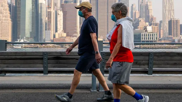An older man and woman wearing masks and walking next to the water with the Manhattan skyline behind them.