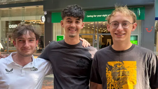 Left to right are England fans James, Nick, Tommy standing in Birmingham high street. James is wearing a white Castore t-shirt and has his left arm around Nick's shoulder. Nick is wearing a dark grey t-shirt while Tommy is wearing a similar colour t-shirt with a yellow box and pattern in the middle of it. All three are smiling and Tommy is wearing glasses.