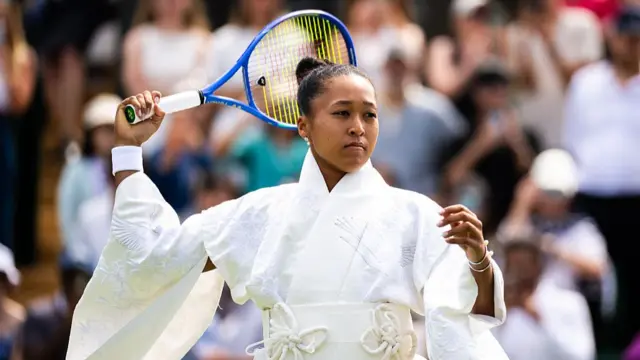 Naomi Osaka, wearing a white kimono and her dark hair in a bun, rehearses a serve her racquet behind her head