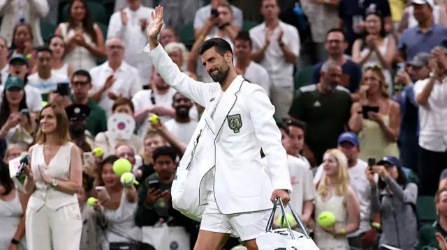 Novak Djokovic, wearing a white blazer and white shorts, carries a white holdall in his left hand and waves his right as he walks off court